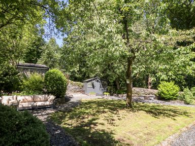A garden with trees a grassy area a gray shed a stone wall and outdoor seating at Yr Hen Ysgol The Old School in Prenteg