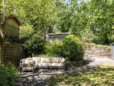 An outdoor garden area with cushioned patio sofa surrounded by trees and bushes at Yr Hen Ysgol The Old School Prenteg