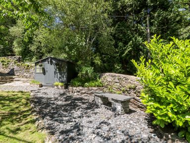 A garden area with a stone bench a grey wooden shed stone walls and trees at Yr Hen Ysgol The Old School in Prenteg