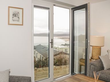 A living room with a wooden table a lamp on a side table gray sofas and glass doors overlooking a coastal view at The Sandy Bee in Benllech