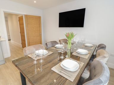 A dining room with a wooden table set with white plates glasses and cutlery and a vase with flowers and wall mounted tv at The Sandy Bee in Benllech