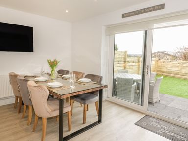 A dining room with a glass table set with plates and glasses and six chairs next to a sliding glass door with outdoor seating at The Sandy Bee in Benllech