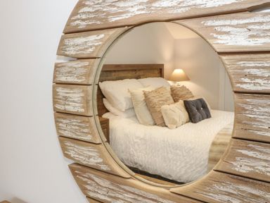 A bedroom with a bed with multiple pillows and a bedside lamp reflected in a round wooden mirror at The Sandy Bee in Benllech