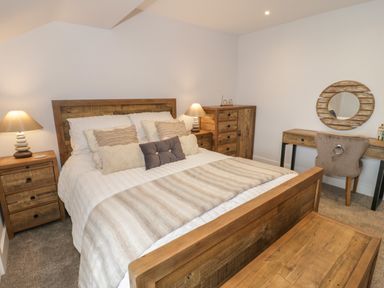 A bedroom with a wooden bed a striped bedspread pillows wooden bedside tables lamps a wooden dresser a desk with a chair and a round mirror at The Sandy Bee in Benllech