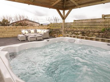 An outdoor area with a hot tub under a wooden roof and grey cushioned seating with cushions near a wooden fence at The Sandy Bee in Benllech