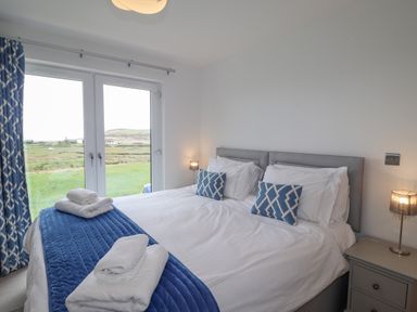 A bedroom with a bed white linens blue pillows and curtains and glass doors showing a grassy landscape at Chandlers Retreat in Newport Pembrokeshire