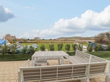 An outdoor patio with white metal benches and table overlooking a grassy field with boats and houses in the distance at Chandlers Retreat in Newport Pembrokeshire