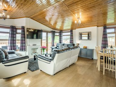 A living room with leather sofas a wall mounted tv a glass coffee table a dining table wooden ceiling and windows with curtains at Lakeview Lodge in South Lakeland Leisure Village