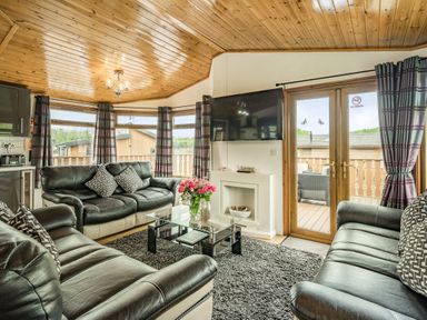 A living room with black leather sofas a glass coffee table a wall-mounted television and glass doors leading outside at Lakeview Lodge in South Lakeland Leisure Village