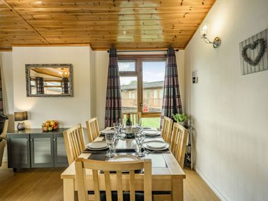 A dining room with a wooden table set for six next to a window with curtains at Lakeview Lodge South Lakeland Leisure Village
