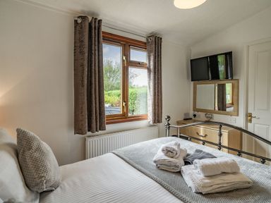 A bedroom with a bed stacked with towels a window with curtains a television mounted above a dresser and a mirror at Lakeview Lodge in South Lakeland Leisure Village