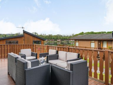 A patio with outdoor wicker furniture and a table with bottles on the deck overlooking wooden cabins and a lake at Lakeview Lodge in South Lakeland Leisure Village