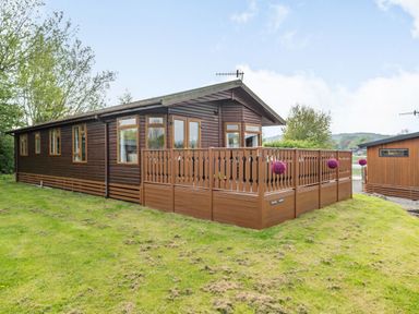 A brown wooden lodge with a fenced porch and hanging flower baskets on a grassy area at Lakeview Lodge in South Lakeland Leisure Village
