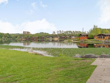 A lake with wooden lodges around it and grass and stone path in the foreground at Lakeview Lodge in South Lakeland Leisure Village