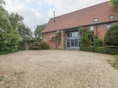A gravel driveway leading to a brick house with a tiled roof and large windows surrounded by bushes and trees at The Barn in Tunstead near Hoveton & Wroxham