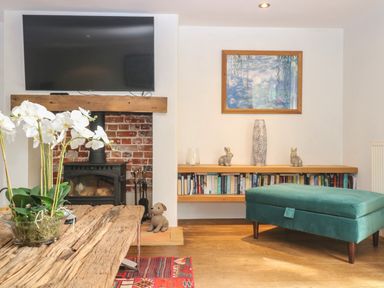 A living room with a wood burning stove under a wall mounted tv a wooden table with flowers and a green upholstered ottoman at The Barn Tunstead near Hoveton & Wroxham