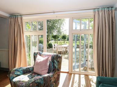 A sitting area with a patterned armchair and a pink cushion in front of glass doors leading to an outdoor patio at The Barn in Tunstead near Hoveton & Wroxham
