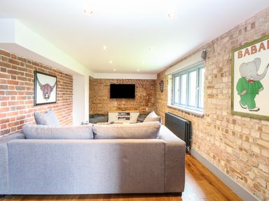 A living room with a grey sofa facing a TV mounted on a brick wall and a window with a radiator below at The Barn in Tunstead near Hoveton & Wroxham
