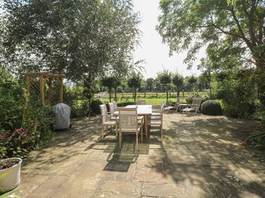 A garden patio with a wooden dining table and chairs surrounded by trees and bushes at The Barn in Tunstead near Hoveton & Wroxham