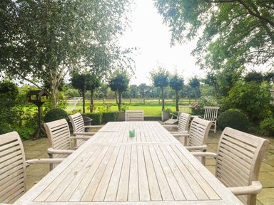 An outdoor patio with a long wooden table and chairs surrounded by trees and a garden at The Barn Tunstead near Hoveton & Wroxham