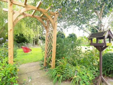 A garden with a wooden archway a bird feeder and a red chair at The Barn Tunstead near Hoveton & Wroxham