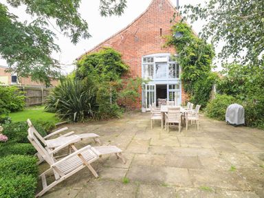 An outdoor patio area with wooden dining table and chairs two lounge chairs and a barbecue grill in a garden at The Barn Tunstead near Hoveton & Wroxham