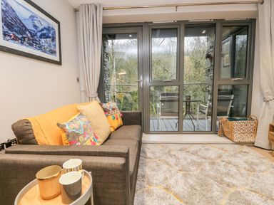 A living room with a sofa and cushions a small round table with cups and candle a large window and glass door leading to a balcony with two chairs and a table at River View in Backbarrow