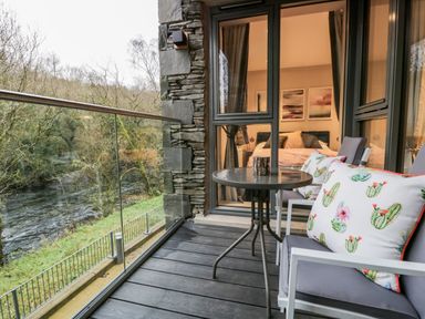 A balcony with chairs a table and cushions overlooking a river with a view into a bedroom at River View in Backbarrow