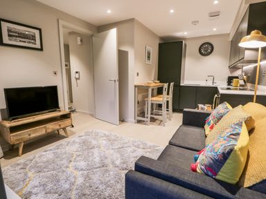 A living area with a gray sofa and pillows a wooden TV stand with a television a small dining table with two chairs and a kitchen at River View in Backbarrow