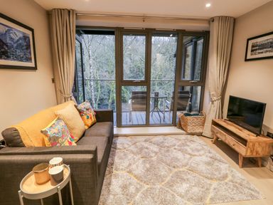 A living room with a gray sofa and patterned cushions next to a coffee table and a wooden TV stand with a television at River View in Backbarrow