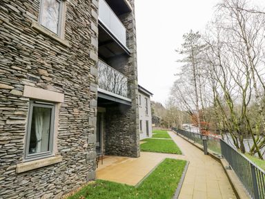 An exterior view of a stone building with balconies beside a paved pathway and trees by a river at River View in Backbarrow