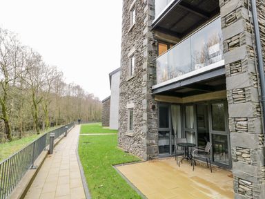 An outdoor patio with table and chairs next to a stone building and a paved path with railing beside grassy areas at River View in Backbarrow