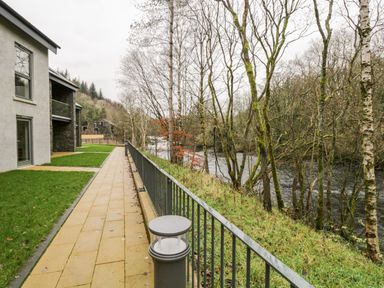 A paved path with grass on one side and a metal railing overlooking a river with trees at River View in Backbarrow