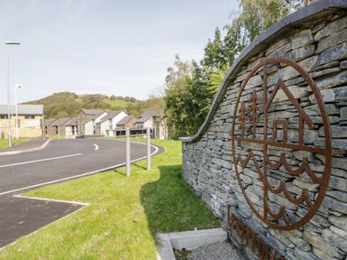 A stone wall with a metal house and tree design beside a curved road with houses and streetlights in the background at Ironworks in Backbarrow