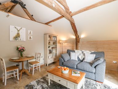 A living room with a gray sofa pillows a wooden coffee table a small dining table with two chairs a bookshelf and wall art at The Barn at Rapps Cottage Rapps near Ilminster