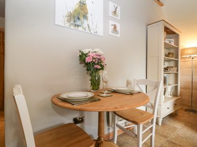 A small dining area with a round wooden table set for two with flowers and plates near a white shelving unit with a lamp at The Barn at Rapps Cottage Rapps near Ilminster