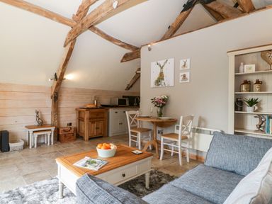 A living room with a gray sofa wooden coffee table bowl of fruit small dining table with two chairs and wooden beams at The Barn at Rapps Cottage in Rapps near Ilminster