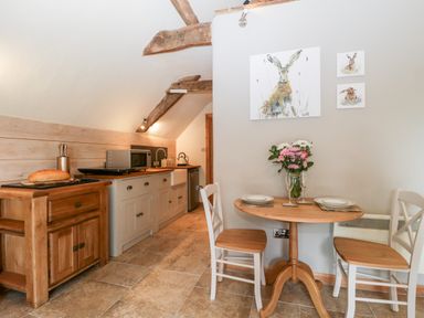 A small kitchen with wooden furniture a table with two chairs and wall art of rabbits at The Barn at Rapps Cottage in Rapps near Ilminster
