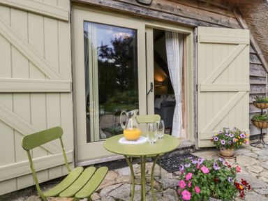 A small green outdoor table and chair set with a jug of orange juice and glasses outside double doors with shutters and flowers on a stone patio at The Barn at Rapps Cottage in Rapps near Ilminster