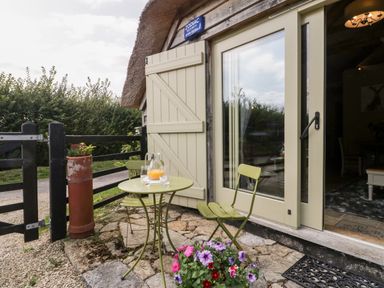 An outdoor patio with a small round table and two chairs next to a glass door with beige shutters at The Barn at Rapps Cottage in Rapps near Ilminster