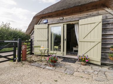 The outside of a cottage with open shutters and a small table with two chairs and flowers at The Barn at Rapps Cottage in Rapps near Ilminster