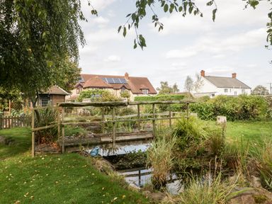 A garden with a wooden bridge over a pond and houses in the background at The Barn at Rapps Cottage in Rapps near Ilminster