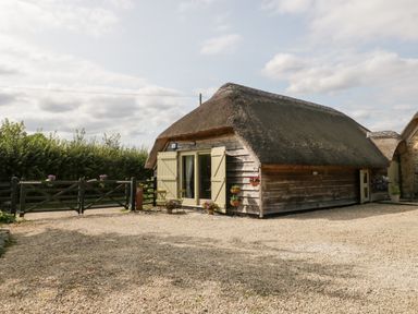 A thatched wooden cottage with a gravel driveway a black gate and outdoor chairs and flowers at The Barn at Rapps Cottage Rapps near Ilminster