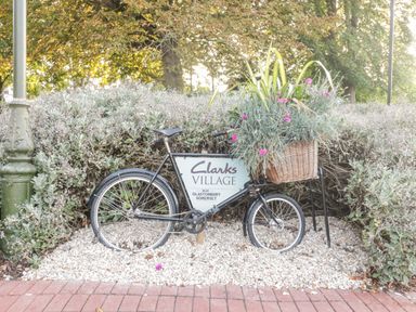 A black bicycle with a basket of flowers and a Clarks Village sign near shrubs and trees at The Barn at Rapps Cottage in Rapps near Ilminster