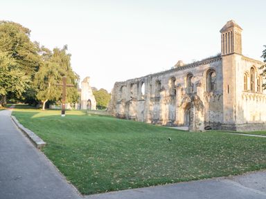 Stone ruins of a large building with arched windows and a tower near a grassy area with a wooden cross and trees at The Barn at Rapps Cottage in Rapps near Ilminster