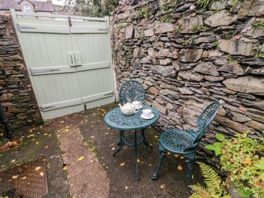 An outdoor patio area with a metal table and two chairs next to a stone wall and a wooden gate at Nutkin Cottage in Bowness-On-Windermere