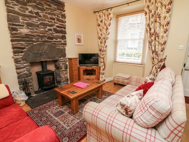A living room with a stone fireplace a wood stove a wooden coffee table a TV a red sofa and a checked sofa at Nutkin Cottage in Bowness-On-Windermere