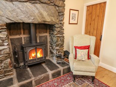A stone fireplace with a wood stove fire next to an armchair with cushions and a wooden door at Nutkin Cottage in Bowness-On-Windermere