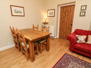 A dining area with a wooden table and six chairs next to a red sofa and wooden door at Nutkin Cottage in Bowness-On-Windermere