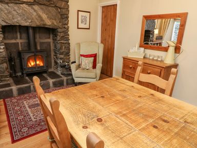 A dining area with a wooden table and chairs next to a stone fireplace and an armchair at Nutkin Cottage in Bowness-On-Windermere